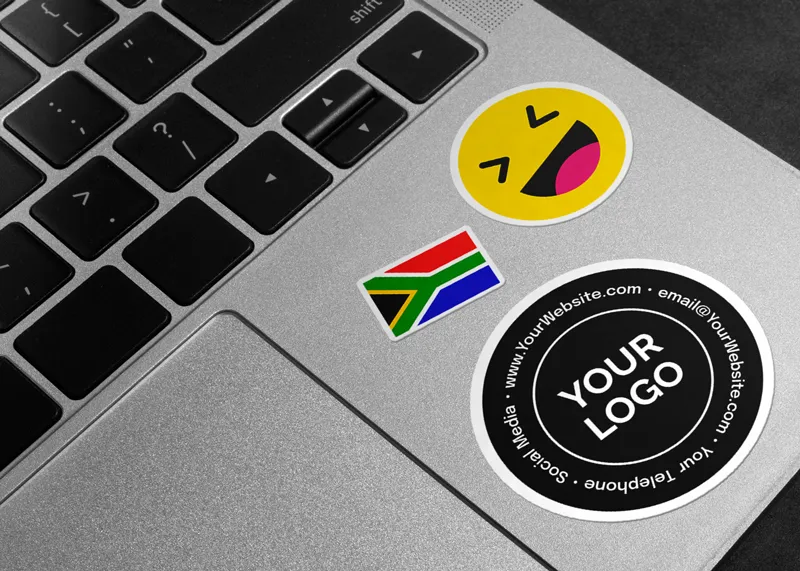 Laptop keyboard with three stickers: a smiling emoji, South African flag, and a round black sticker labeled "YOUR LOGO" with placeholder text
