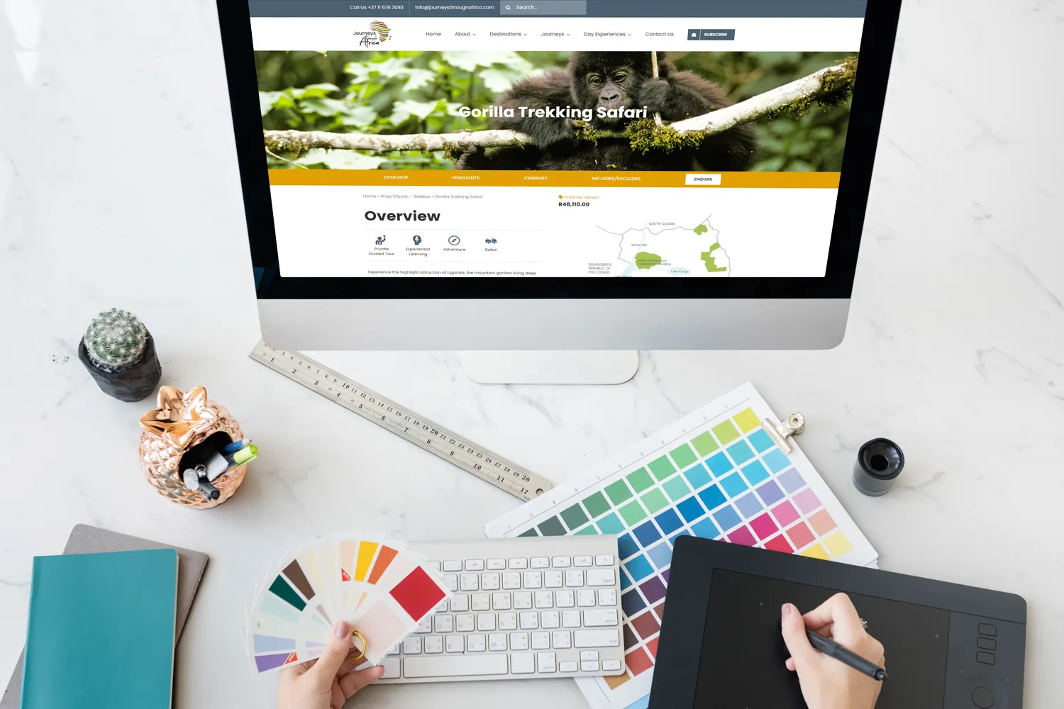 A desktop displays a custom design for a "Gorilla Trekking Safari" site. Nearby, a person holds a color swatch, with design tools and color samples on a marble desk.